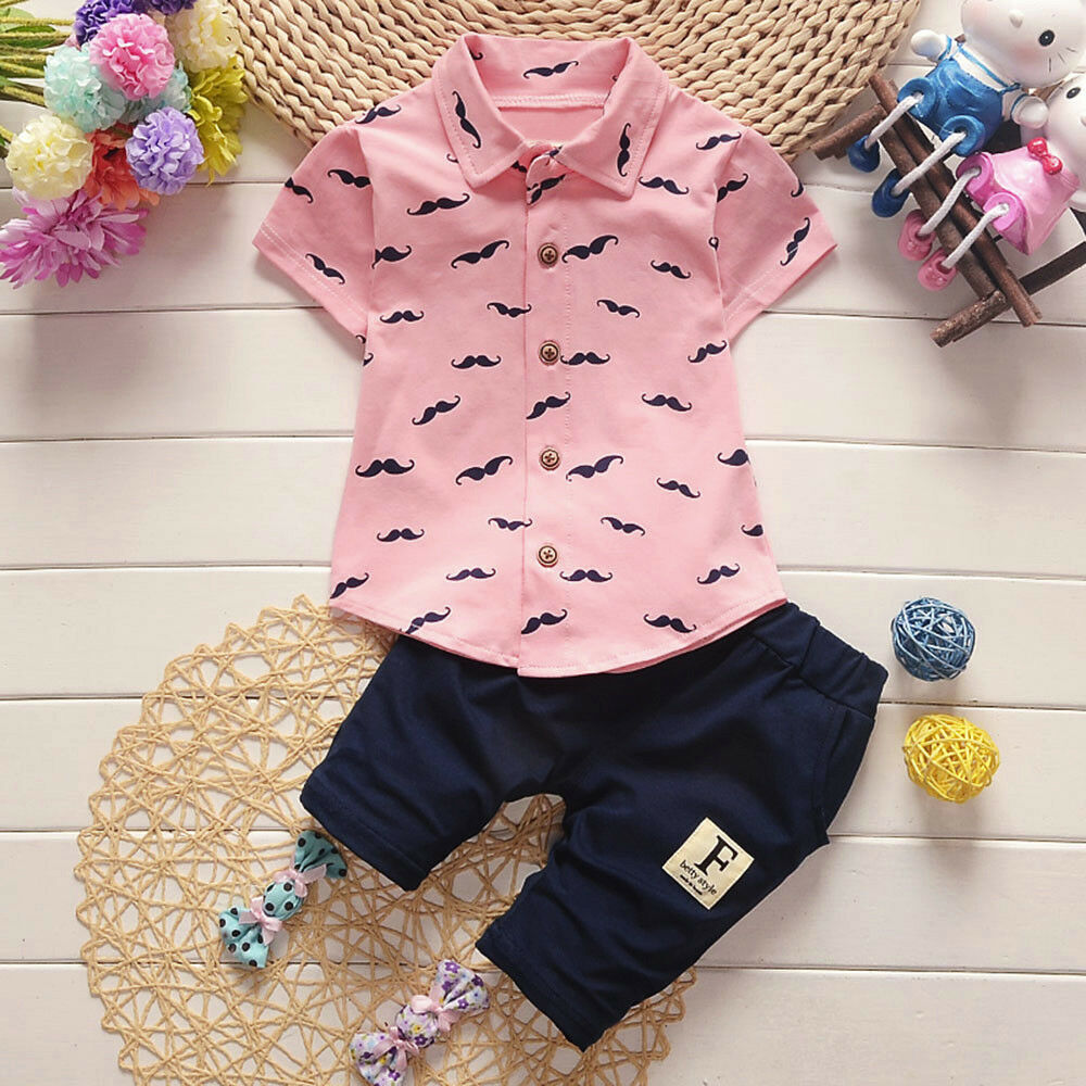 Children's outfit with pink shirt and navy shorts on a wooden surface with toys and flowers.