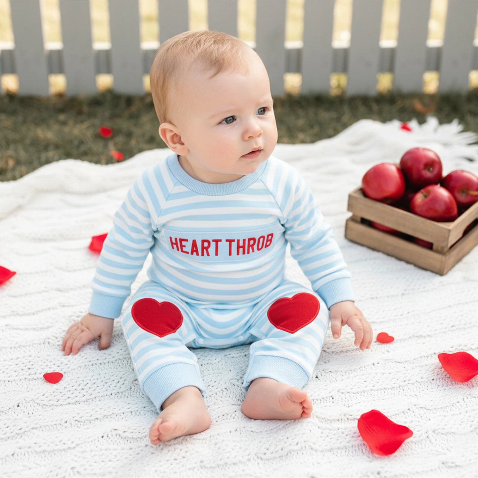 Baby in a 'HEART THROB' onesie surrounded by red petals and apples on a blanket.