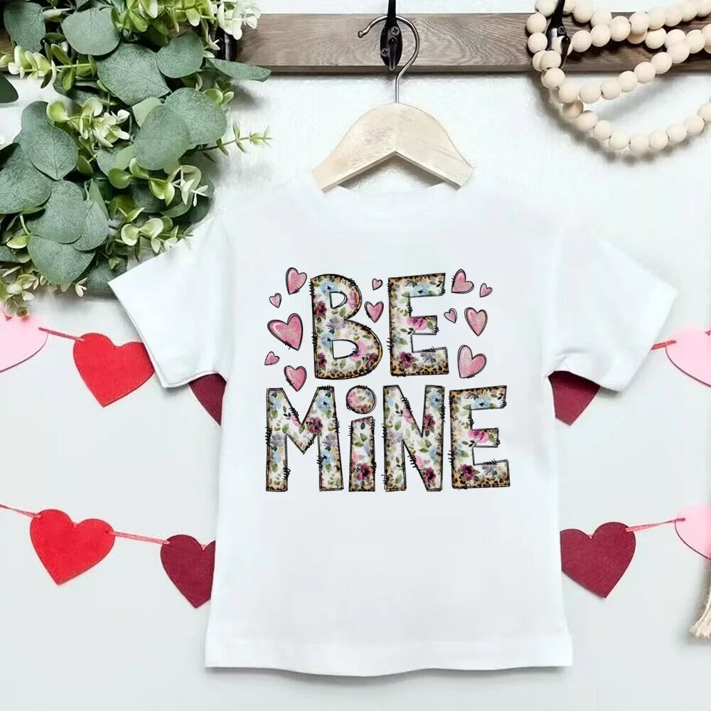White t-shirt with 'BE MINE' text and hearts on a hanger with decorative elements.
