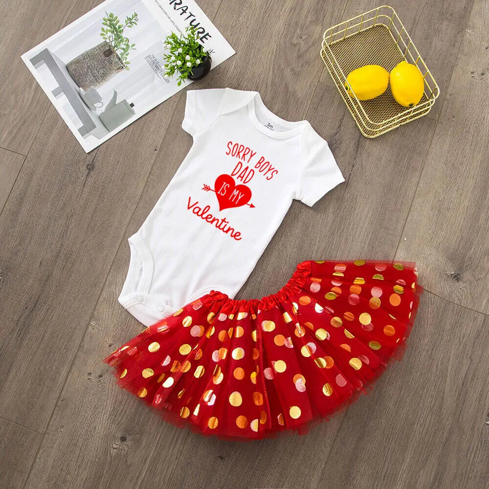 White baby onesie with red text and a red polka dot skirt on a wooden floor.