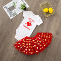 White baby onesie with red text and a red polka dot skirt on a wooden floor.