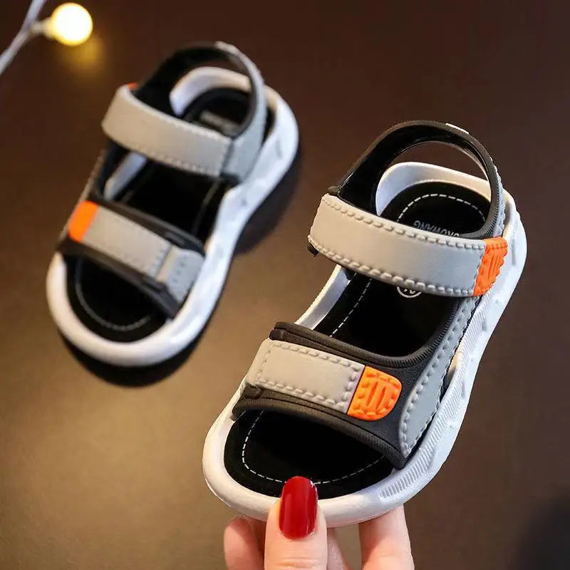 Children's sandals with gray and black straps held by a hand with red nail polish on a brown background.