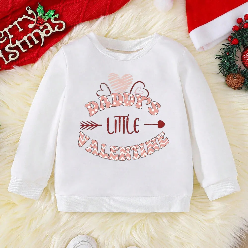 White sweatshirt with 'Daddy's Little Valentine' text on a textured surface with Christmas decorations.