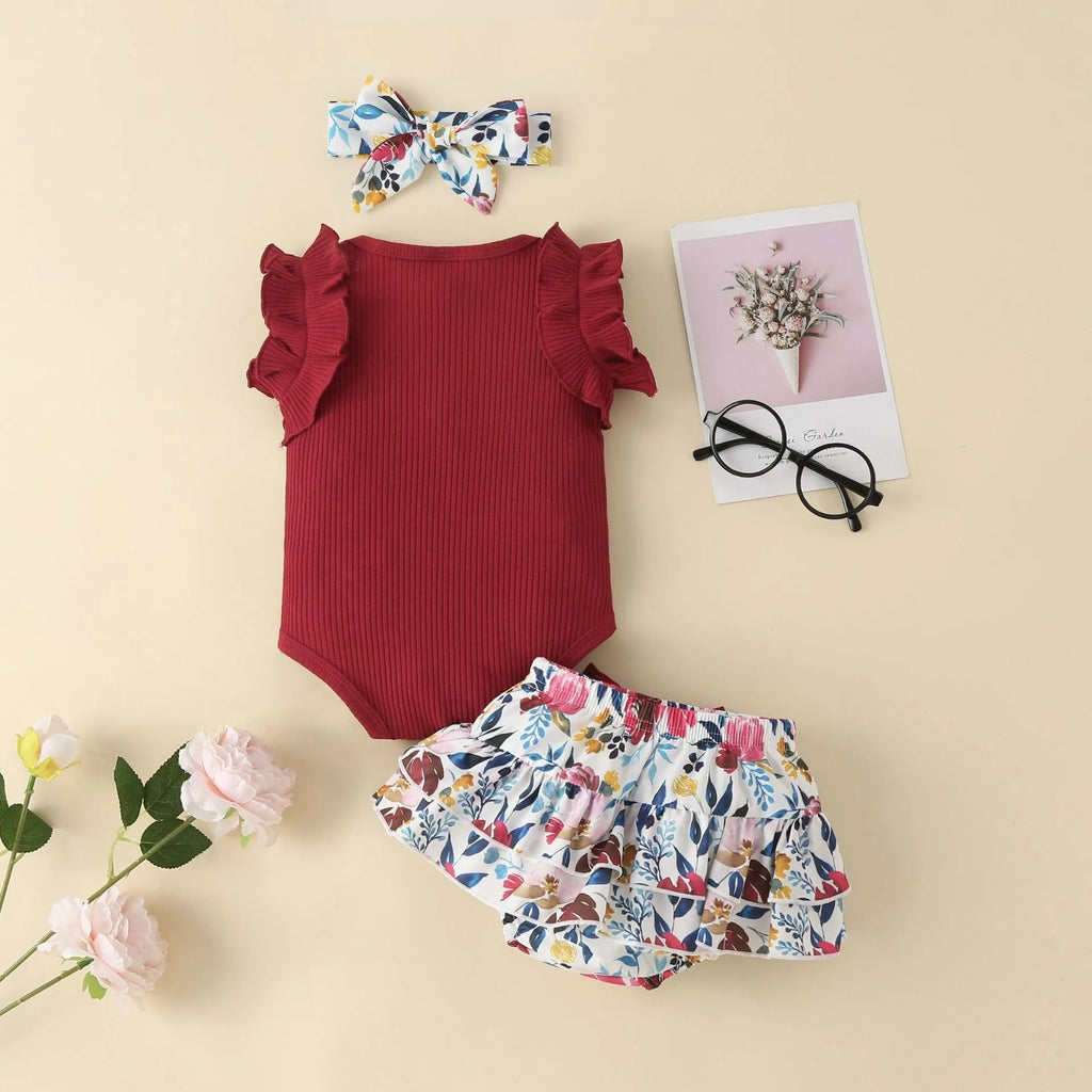 Baby outfit with red onesie, floral bloomers, and headband on a beige background