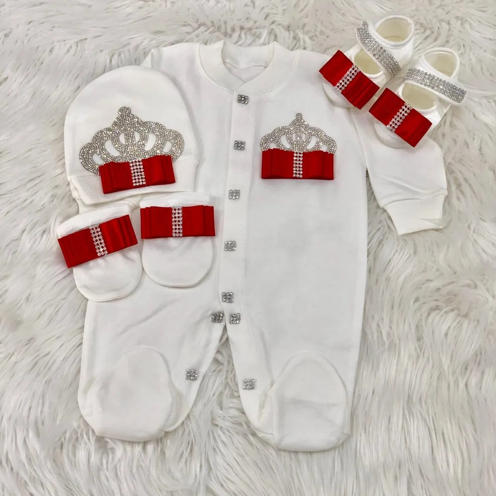 Baby outfit set with white onesie, hat, and booties featuring red bows and silver crowns on a light gray background.