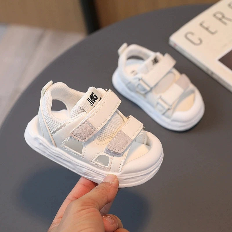 White children's sandals with velcro straps held by a hand on a dark surface.