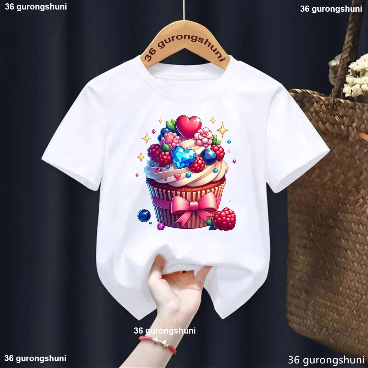 White t-shirt with a colorful cupcake design on a hanger against a dark background.