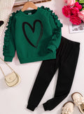 Green sweater with a heart design and black pants on a white surface.