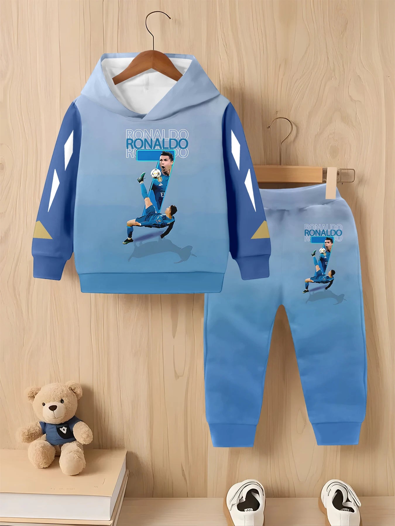 Children's blue Ronaldo-themed hoodie and pants set on a wooden background with a teddy bear and books.
