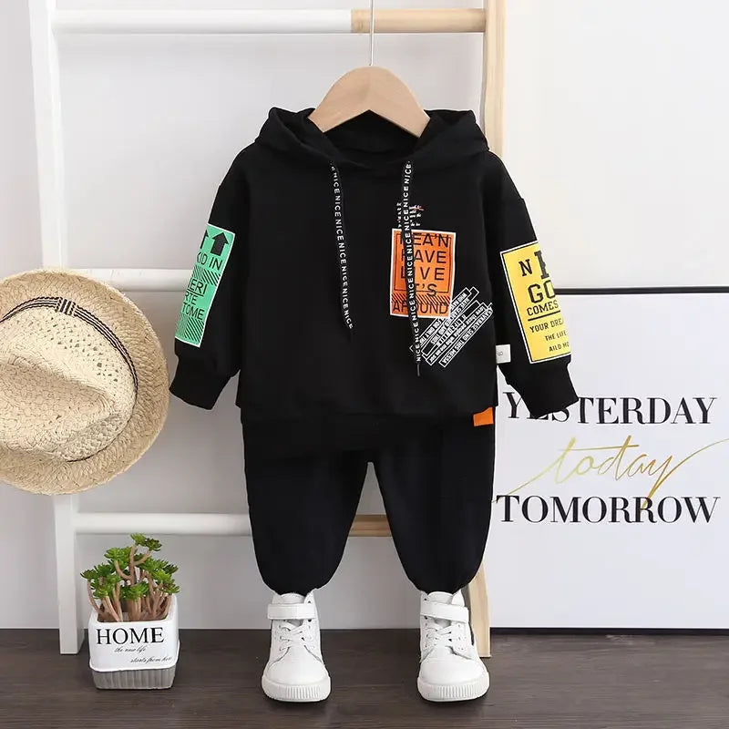 Black hoodie and pants set with colorful patches on a hanger, next to a hat and plant.