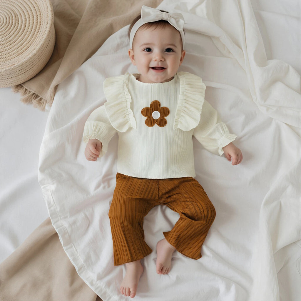 Baby wearing a white sweater with a brown flower design and brown pants, lying on a white blanket.