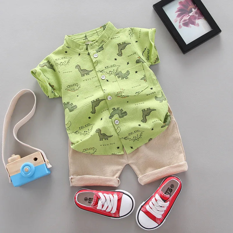 Children's green shirt with dinosaur pattern and beige shorts on a gray surface with red sneakers and a toy camera.