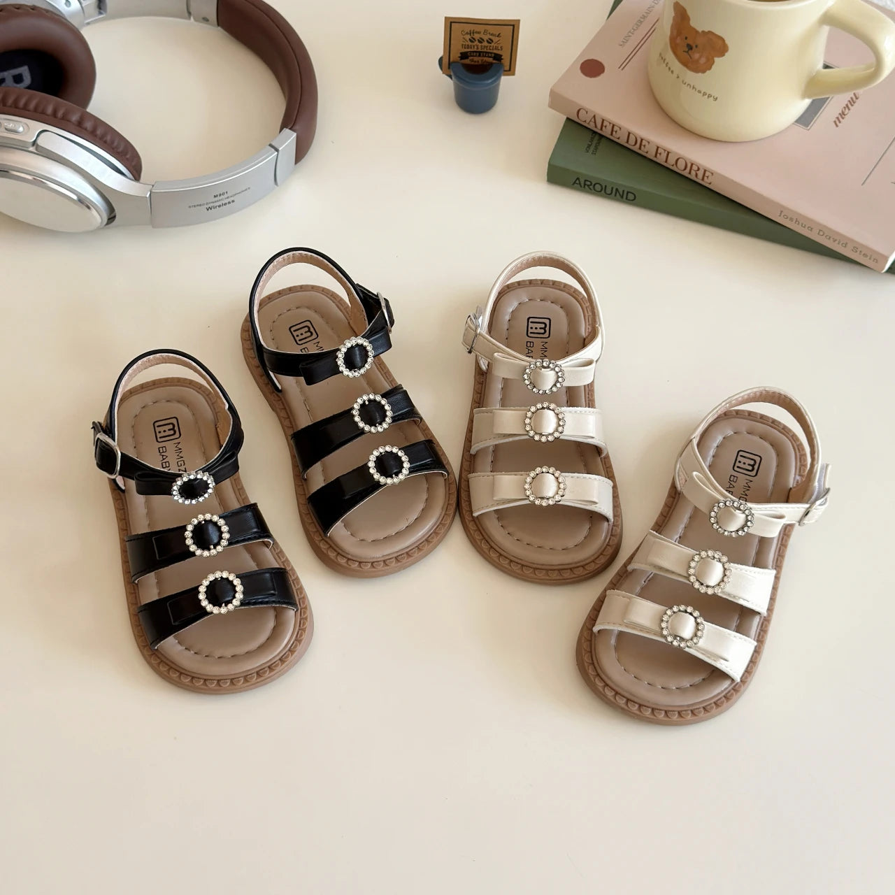 Pair of children's sandals on a light surface with books and a mug in the background.