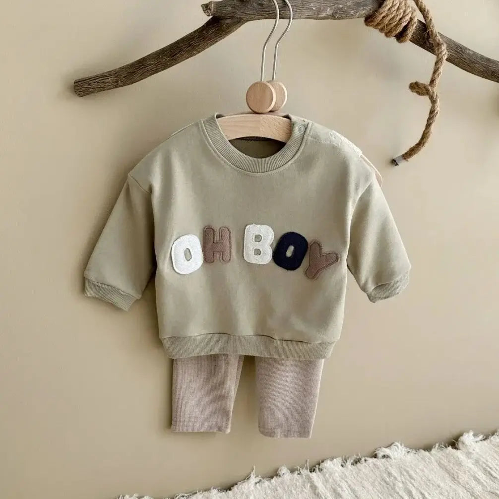 Children's outfit with 'OH BOY' text on a wooden hanger against a beige wall.