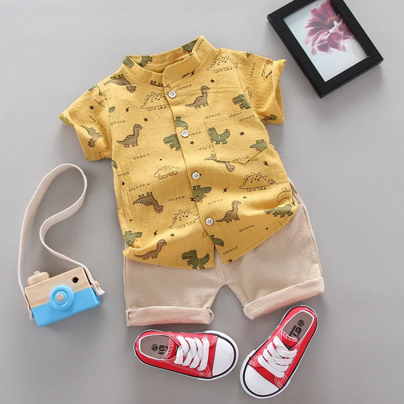 Children's yellow shirt with dinosaur pattern and beige shorts on a gray surface, with red sneakers and a toy camera.
