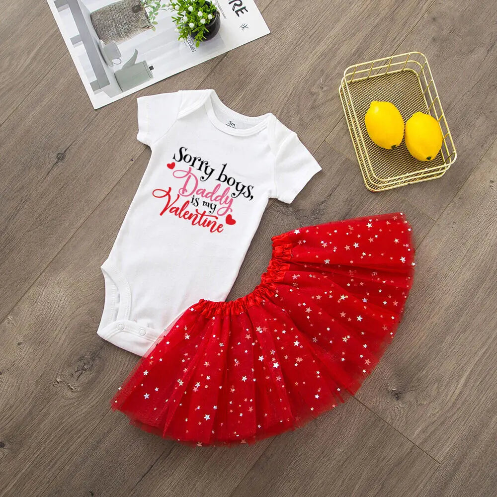 White baby onesie with text and red tutu skirt on a wooden floor.