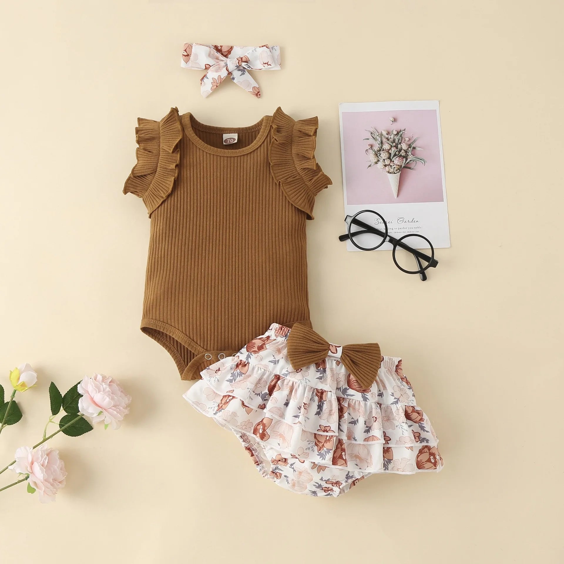 Baby outfit set with brown romper, floral shorts, headband, and glasses on a beige background.
