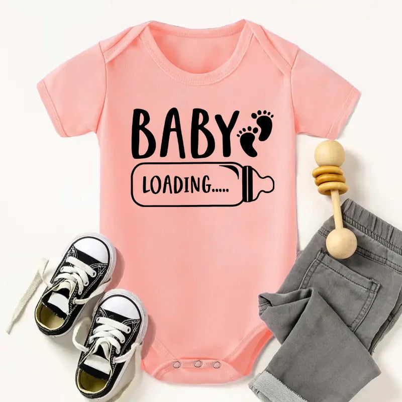 Pink baby onesie with 'Baby Loading' text, black sneakers, gray pants, and wooden rattle on a white background
