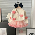 Children's outfit with a white cardigan with pink heart patterns and a pink skirt, displayed on a hanger.