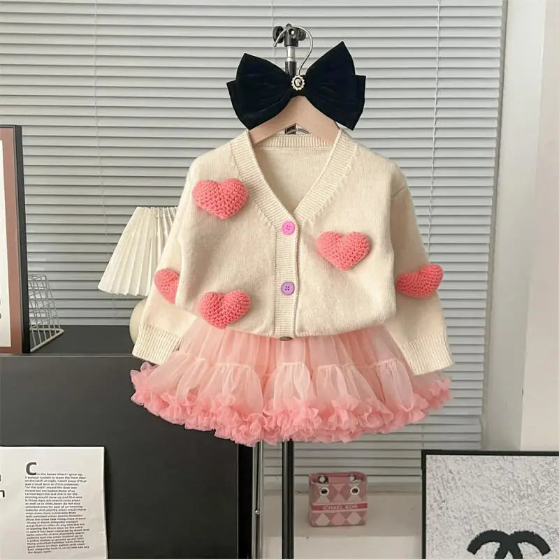 Children's outfit with a white cardigan with pink heart patterns and a pink skirt, displayed on a hanger.