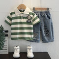 Children's green and white striped polo shirt with a graphic, blue jeans, and white sneakers on a wooden hanger against a white wall.