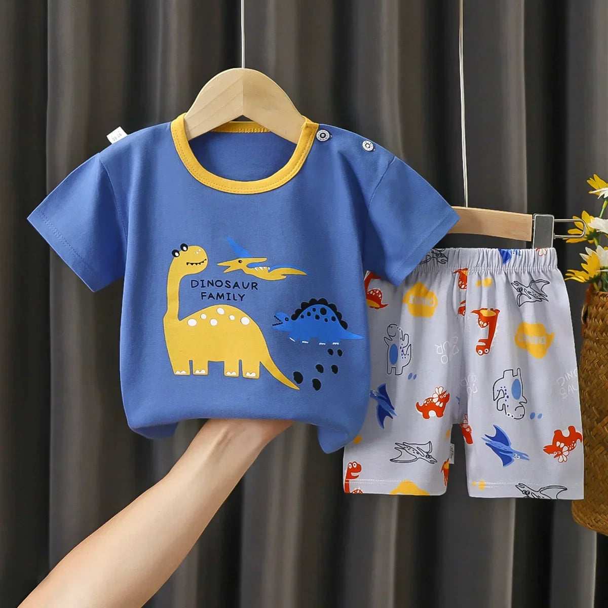 Children's blue t-shirt with dinosaur design and patterned shorts on a hanger against a dark curtain background.