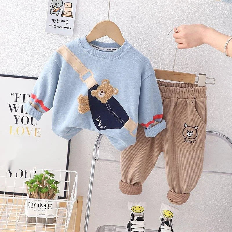 Children's blue sweater with bear design and brown pants on hangers against a light gray wall.
