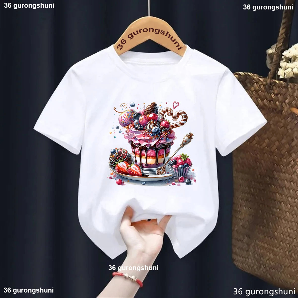 White t-shirt with a colorful cupcake design on a hanger against a dark background.