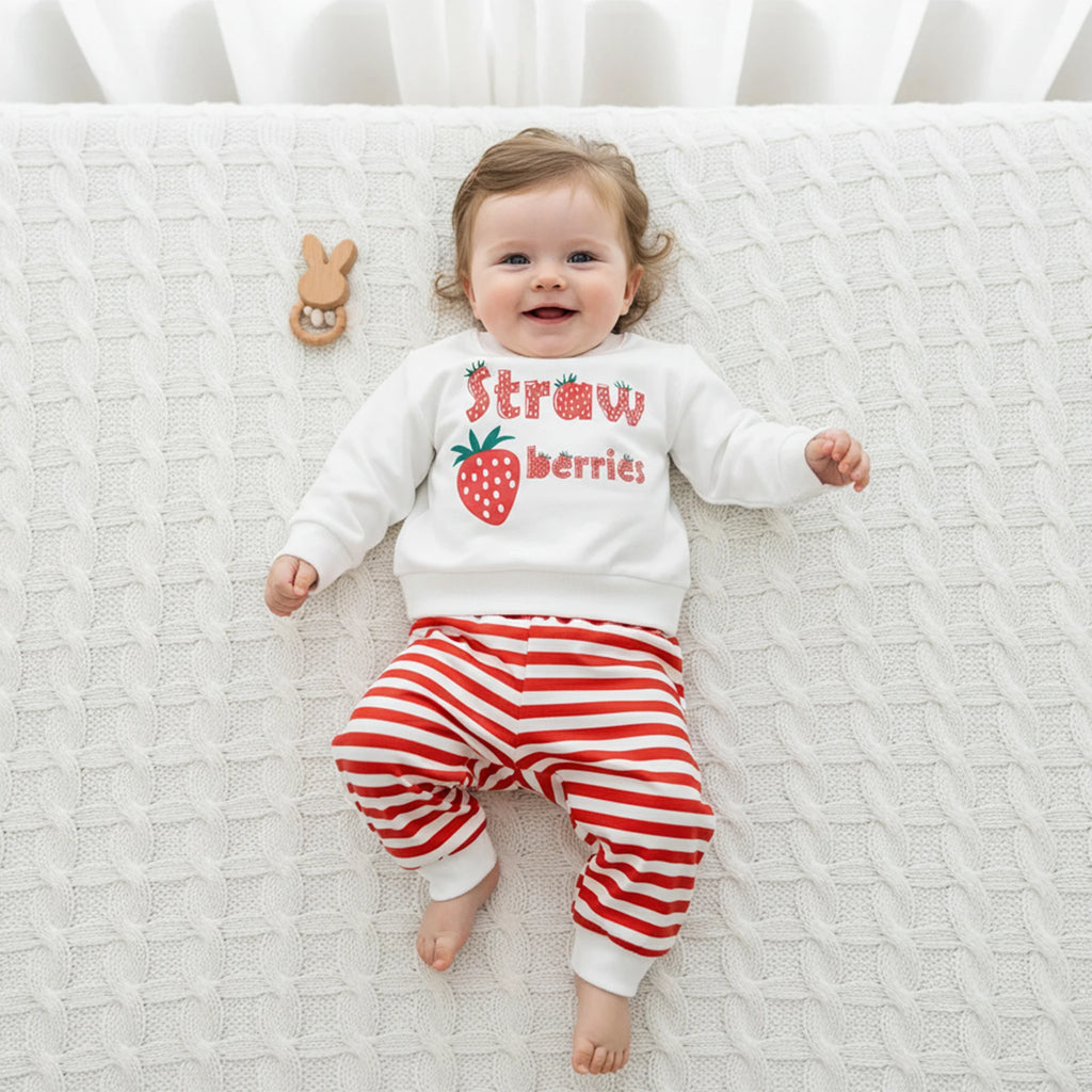 Baby wearing a white sweatshirt with 'Strawberries' text and red striped pants, lying on a textured white surface.