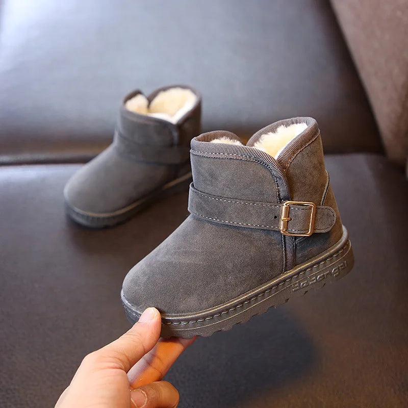 Gray children's snow boots with a buckle held by a hand on a dark surface.