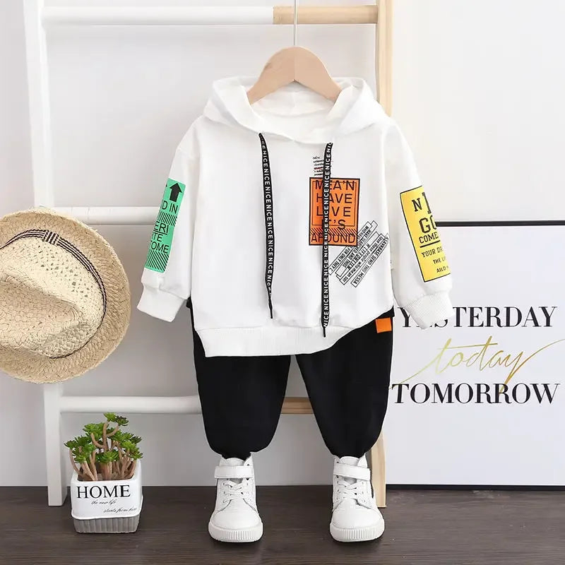 White hoodie with colorful text and graphics, black pants, white sneakers on a wooden floor with a hat and plant in the background.
