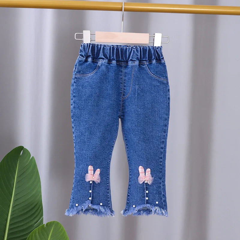 Blue denim pants with bow details on a hanger against a gray background