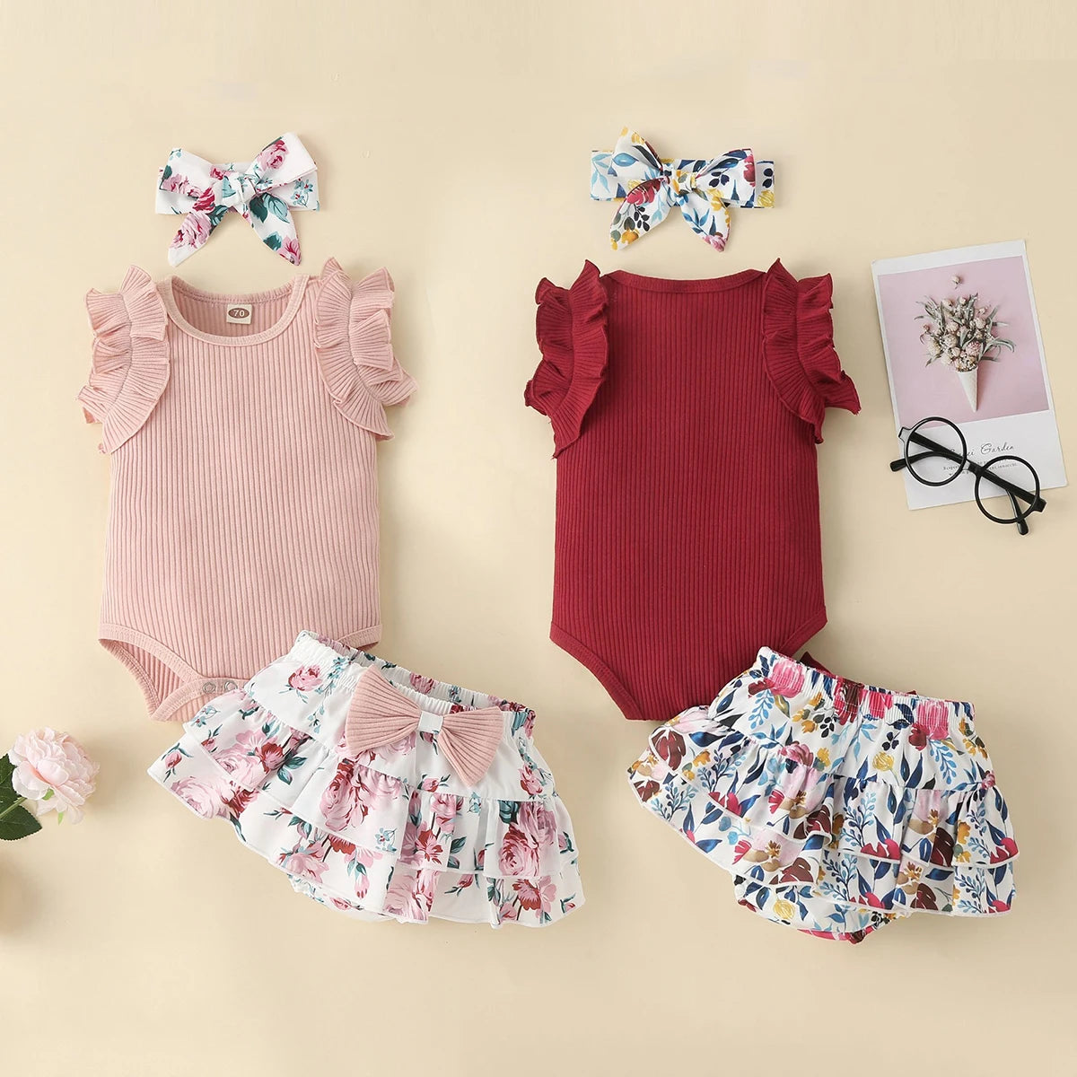Children's outfit set with pink and red rompers, floral shorts, and headbands on a beige background.