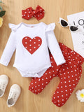 White baby romper with red heart design, red polka dot pants, white shoes, and a red bow on a wooden surface.