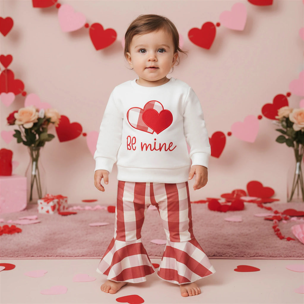 Child wearing a Valentine's Day-themed outfit with a heart design and 'bf mine' text, standing in a decorated room with heart decorations.