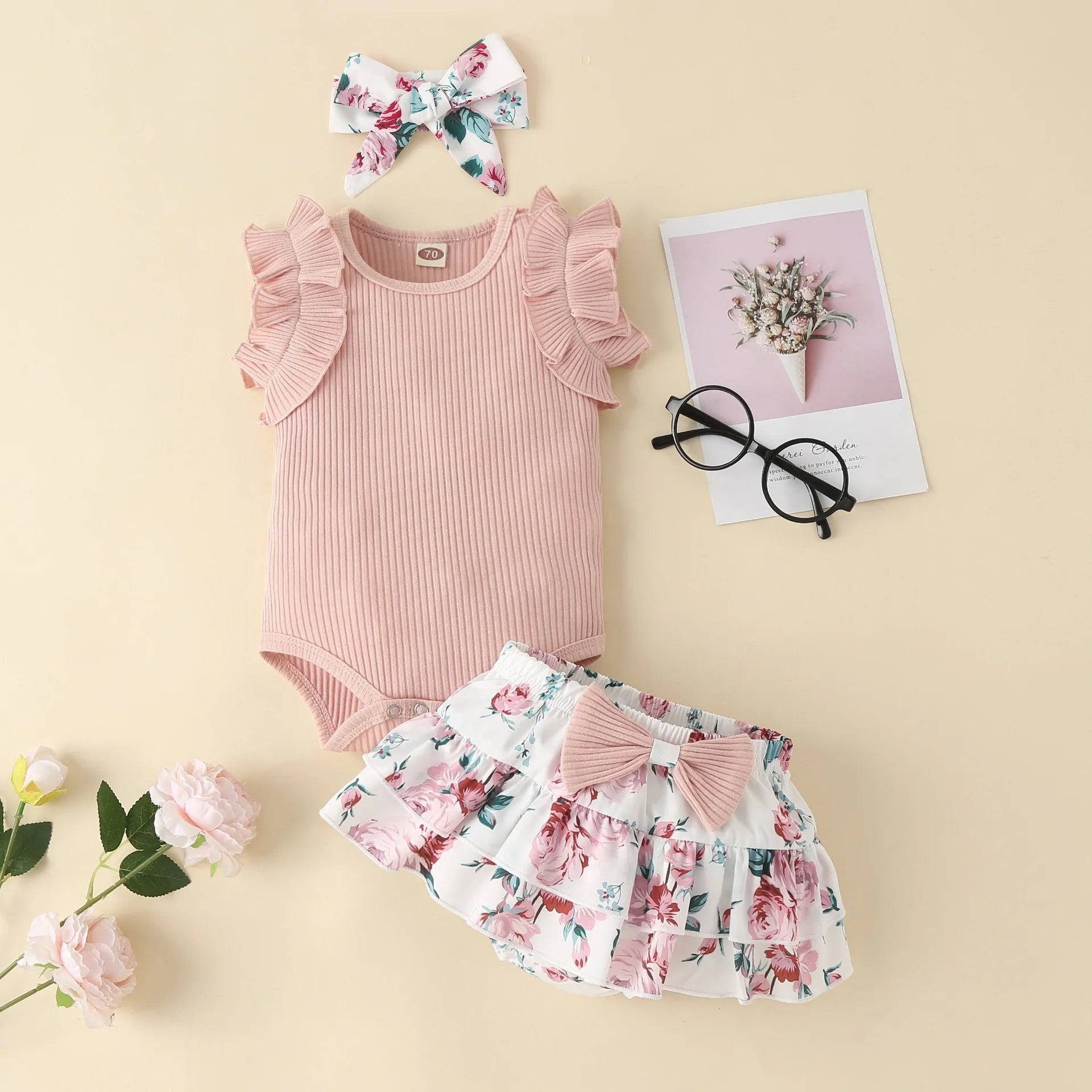 Baby outfit with pink romper, floral shorts, and headband on a beige background